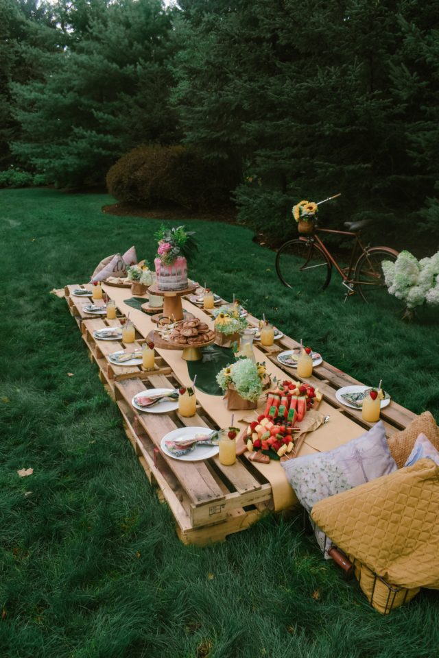Photo of several wooden pallets on a grassy lawn. The pallets are set like a table, with plates, knives, drinks, and produce laid out for a meal. There is a bicycle in the background