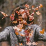 In focus: fall leaves floating through the air in the foreground. Out of focus: A person with dark skin, dark hair and a grey sweater, with arms spread wide, is smiling in the background