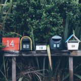A series mailboxes of different shapes and colors, sit side-by-side in front of greenery.