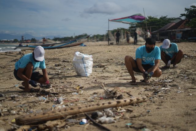 Three dark-skinned people squatting to pick up trash on a beach. They all wear light blue t-shirts, and two have white baseball caps. A boat and a few buildings, trees and people are visible in the background
